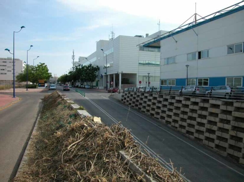 Fotografía de una calle con edificios industriales, coches estacionados y un muro de contención.