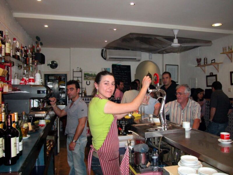 Una camarera en un bar, con una botella de licor en la mano izquierda y una taza de café en la derecha, mientras atiende a clientes. El ambiente es casual con una barra de madera y estantes llenos de bebidas alcohólicas.