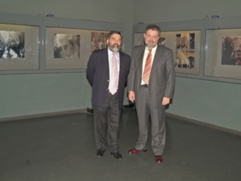 Dos hombres en trajes posan frente a una pared con varias fotografías.