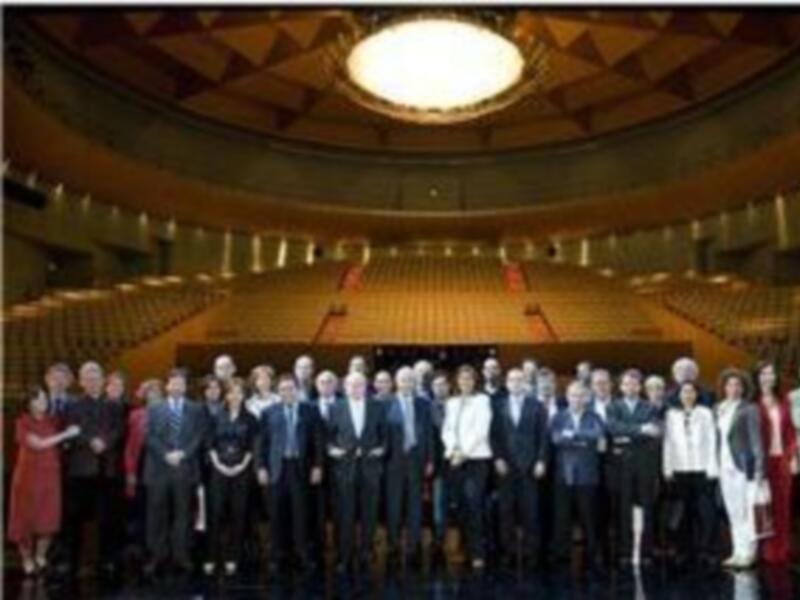 Una reunión formal en un auditorio con una iluminación dorada y luces de techo. Los asistentes están vestidos formalmente, posando para una foto en el centro del escenario.
