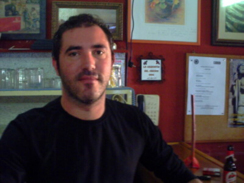 Un hombre con barba y bigote, mirando a la cámara. Detrás de él, una pared con carteles y un calendario. A la derecha, una botella de cerveza.