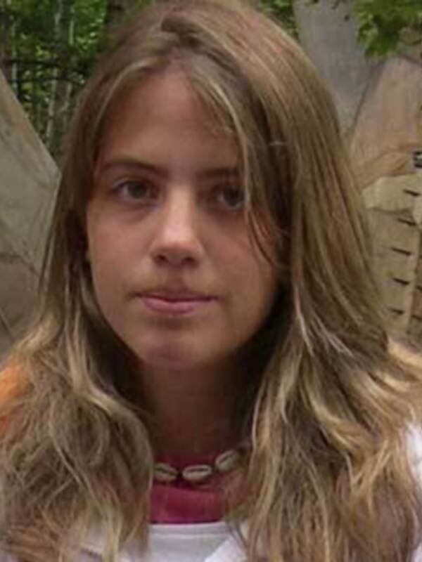 Una persona con cabello largo y ondulado, usando una camiseta blanca y una collana con cuentas, frente a un fondo de madera con forma de animal.