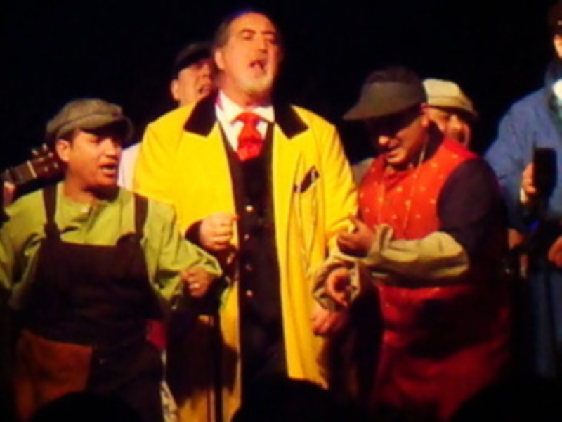 Una escena teatral con un hombre en el centro, vestido con una chaqueta amarilla y corbata roja, cantando emocionalmente. A su alrededor, otros personajes con trajes de trabajo y sombreros observan la actuación. La iluminación es dramática, enfocándose en el cantante principal.