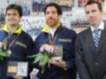 Dos atletas posan con medallas y flores, uno de ellos sostiene un trofeo. Detrás de ellos, se observa una pared con fotos y información deportiva. A la derecha, un hombre en traje sostiene una bandera europea.