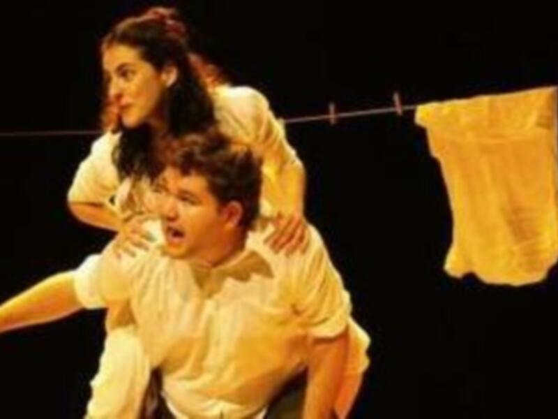 Dos personas en una escena teatral, uno apuntando hacia la otra con un fondo de ropa colgada.