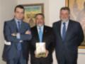 Tres hombres en trajes posan con un libro titulado 'Los Exploradores de Sevilla' en un entorno formal.