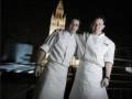 Dos chefs en uniforme blanco posan frente a un edificio histórico de noche.