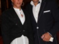Dos personas posando en un evento formal. La persona de la izquierda lleva una chaqueta negra con detalles blancos y pantalones negros. La persona de la derecha está vestida con un traje negro y una camisa blanca. Ambos están sonriendo y parecen estar en un ambiente de celebración o gala.