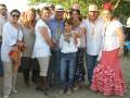 Una familia feliz en un ambiente festivo, con una mujer en falda de volantes roja y blusa blanca, otro en sombrero beige y camisa azul con amarillo, y una niña en jeans y chaqueta blanca.