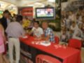 Un hombre en camisa blanca y pantalones vaqueros está firmando autógrafos para una persona en un vestido rosa. En la mesa hay libros y regalos. Al fondo, una pantalla muestra un partido de fútbol mientras otros asistentes observan.