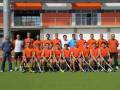 Un equipo de hockey sobre césped posa en un campo de juego, todos vestidos con camisetas naranjas y pantalones cortos. Al fondo, se pueden ver los muros de un edificio con ventanas y una barandilla.