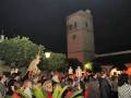Una procesión nocturna con una imagen religiosa en el centro, rodeada de personas y una torre de la iglesia al fondo.