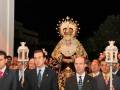 Fotografía nocturna de una procesión religiosa en España, con una imagen venerada en el centro y varios hombres en trajes de gala a su alrededor, sosteniendo estandartes.