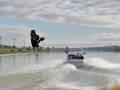 Un wakeboarder realiza un salto impresionante sobre el agua mientras un barco lo guía. El cielo azul con nubes dispersas y la orilla de la ciudad en el fondo.