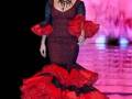 Una modelo desfilando en un vestido flamenco rojo con detalles negros y una falda larga con volantes. La imagen destaca la elegancia y tradición de la moda flamenca.