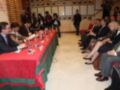 Una reunión formal en una sala con paredes de madera y carteles. Una mesa cubierta con mantel rojo-verde, con personas sentadas y otros en pie. La atmósfera es seria, probablemente una conferencia o negociación.
