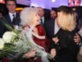 Una anciana con un ramo de flores blancas se abraza con una mujer joven. Detrás, hombres en trajes conversan entre sí. La escena parece ser una celebración o evento formal.