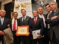 Un grupo de hombres en trajes posa con un certificado y un cartel promocional de eventos taurinos en Andalucía.