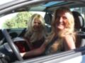 Dos mujeres con cabello largo y rubio están sentadas en un automóvil, una de ellas sonríe ampliamente mientras la otra mira hacia el lado. La imagen sugiere una escena cómica o divertida, posiblemente relacionada con el humor o la comedia.