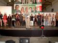 Una ceremonia formal con varios hombres y mujeres en trajes, posando frente a un póster de campaña política. La escena está ambientada en una sala con iluminación artificial y un techo de madera.
