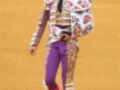 Un torero en traje tradicional de color naranja y blanco con detalles florales, camisa blanca con estampado floral, pantalones morados y guantes negros. El fondo es un terreno de toros amarillo.