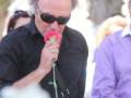Un hombre con gafas negras y una camisa negra, sosteniendo un ramo de flores rojas. La imagen parece ser de una ceremonia o evento con personas en el fondo, posiblemente un funeral.