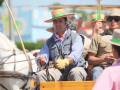 Un hombre en un traje de carruajes, con una corbata y guantes amarillos, montando un caballo blanco en una feria o evento. Detrás de él, hay personas con sombreros y vestimenta tradicional.