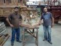 Dos artesanos posan junto a un mueble tallado con detalles ornamentales en una tienda de muebles de madera tallada.