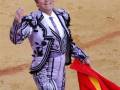 El torero, vestido con un traje tradicional de color púrpura y blanco, ondea una bandera española mientras se encuentra en un anfiteatro de arena.