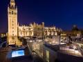 La Giralda, la catedral de Sevilla y otros edificios históricos se destacan en esta imagen nocturna. La torre Giralda, una de las más altas de España, se ilumina en el cielo azul oscuro. En primer plano, un balcón con una piscina iluminada ofrece una perspectiva única de la arquitectura y el paisaje urbano.