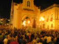 Fotografía nocturna de una procesión religiosa en un lugar histórico, con una gran multitud de personas observando y participando. La imagen muestra una estatua monumental en el centro, rodeada por flores y luces. El fondo es una arquitectura de estilo colonial, con detalles dorados y ventanas. La noche iluminada por farolas y luces de la procesión, creando un ambiente festivo y religioso.