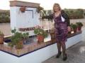 Una mujer con un vestido de leopardo posa frente a una terraza con plantas y flores. La imagen muestra un ambiente tranquilo en un entorno rural o mediterráneo, con una casa blanca y terracota.