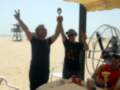 Dos hombres celebran en la playa, uno levanta una copa y el otro sostiene un trofeo. En segundo plano, se puede ver un helicóptero y una torre de vigilancia.
