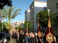 Una procesión religiosa con personas vestidas de negro, banderas y cruces en el camino. Edificios al fondo con balcones y palmeras.