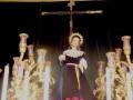 Una figura vestida con un traje oscuro y dorado, posiblemente una imagen religiosa o de la Virgen, está frente a un crucifijo en un entorno oscuro con velas doradas y rojas.