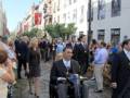 Un hombre en una silla de ruedas se dirige por una calle con edificios blancos y ventanas. La gente lo observa mientras camina hacia el centro de la imagen, donde hay una multitud.