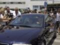 Un hombre se dirige hacia un coche azul, rodeado por una multitud de personas con cámaras y teléfonos. La escena parece ser en una calle con edificios al fondo, posiblemente durante un evento o protesta.
