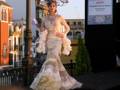 Una modelo presenta un vestido de gala blanco y dorado con detalles florales, frente a una torre y una pancarta que anuncia un evento en Sevilla.