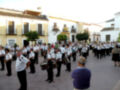 Banda de música desfilando en una plaza de pueblo con edificios blancos y balcones.