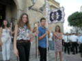 Una joven sonríe mientras sostiene una bandera con un escudo y una cruz, acompañada por otros jóvenes en una procesión. La imagen muestra una calle con edificios históricos y un cielo claro, probablemente durante una celebración o fiesta.