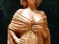 Esta es una estatua de una mujer con cabello largo y ondulado, vestida con un atuendo tradicional que incluye una capa larga y un pañuelo en el hombro. La escultura parece ser de una época antigua, posiblemente romana o griega.