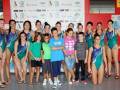 Una foto de un grupo de niñas y niños posando frente a una pared con varios logotipos. Las niñas están vestidas con uniformes de gimnasia y los niños con camisetas y shorts. La pared tiene un fondo blanco con logos de diferentes organizaciones deportivas.