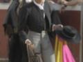 Un torero se prepara para el toro, con un caballo negro detrás. El torero lleva una chaqueta negra y pantalones grises, con un sombrero negro. El caballo tiene una capa amarilla y rosa en el fondo, con la arena de la plaza de toros visible.