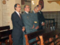 Three men, two in military uniforms and one in a suit, standing in a room with a decorative wall panel and a wooden bench.