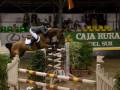 Un jinete monta un caballo en una competencia de salto, saltando sobre un obstáculo blanco y rojo. La imagen se enfoca en el momento de la salto, con un público observando desde los stands. En el fondo, se puede ver una publicidad de "CAJA RURAL DEL SUR".