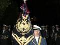 Un hombre en uniforme militar, posiblemente de la policía o el ejército, sostiene un bastón con una bandera y una estatua en la parte superior. La escena parece ser nocturna, con luces de fondo que sugieren una ceremonia o desfile.