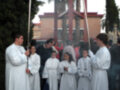 Una procesión religiosa en un entorno urbano con personas vestidas de blanco y otros en trajes formales. Hay palmeras y edificios al fondo, con un cielo despejado.