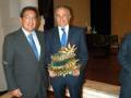 Dos hombres en trajes posan con un trofeo dorado y verde, en un entorno formal.