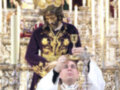 Imagen de un sacerdote en vestimenta religiosa, posiblemente durante una celebración religiosa o procesión. El sacerdote sostiene un objeto de culto en su mano izquierda, mientras que la derecha parece estar en actitud de bendecir o dar un gesto de protección. La vestimenta es elaborada, con detalles dorados y una capilla que cubre la cabeza. Al fondo se observa un altar con adornos y figuras de oro, lo que sugiere una escena de importancia religiosa o ceremonial.