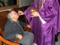 Un sacerdote en vestido púrpura bendice a una anciana sentada en un sillón.